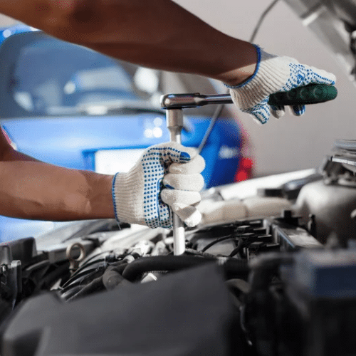 Person wearing gloves uses a ratchet tool to work on a car engine, performing Mazda maintenance with a blue car visible in the background.