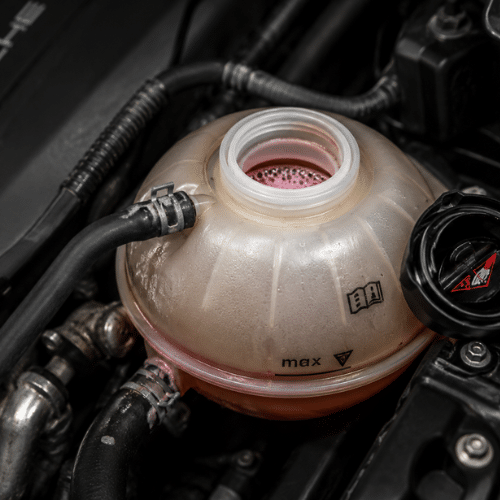 A close-up of a car’s coolant reservoir, partially filled with pink coolant, with the cap removed and visible hoses attached.