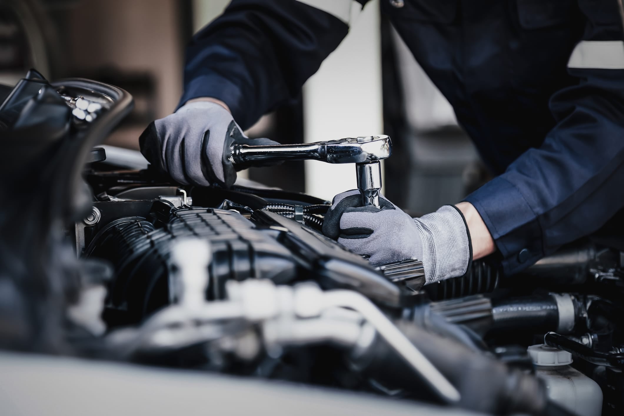 A mechanic wearing gloves uses a ratchet wrench to work on a car engine in a workshop.