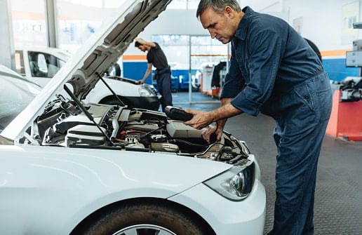 A mechanic in a blue jumpsuit inspects the engine of a white car with the hood open inside an auto repair shop.
