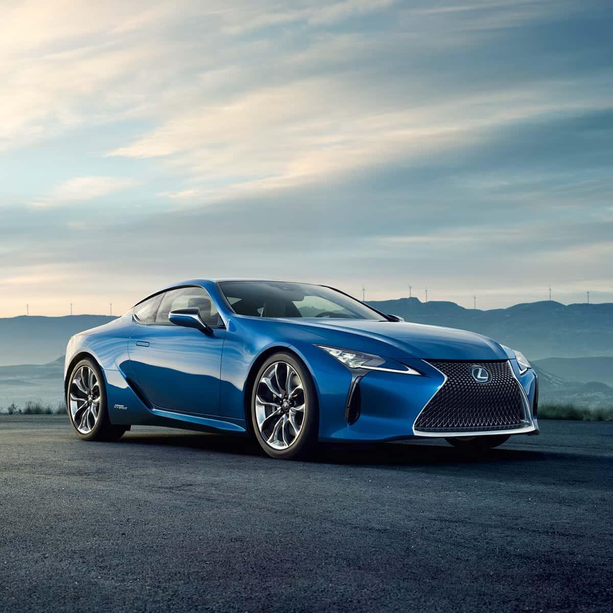 A blue Lexus coupe, well-kept with regular Lexus maintenance, is parked on an empty road near South Jordan, with mountains and a cloudy sky in the background.