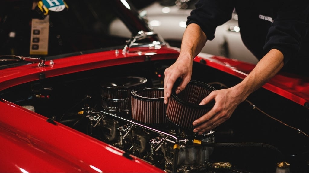 A person’s hands are working under the hood of a red car, adjusting or installing a round engine air filter.