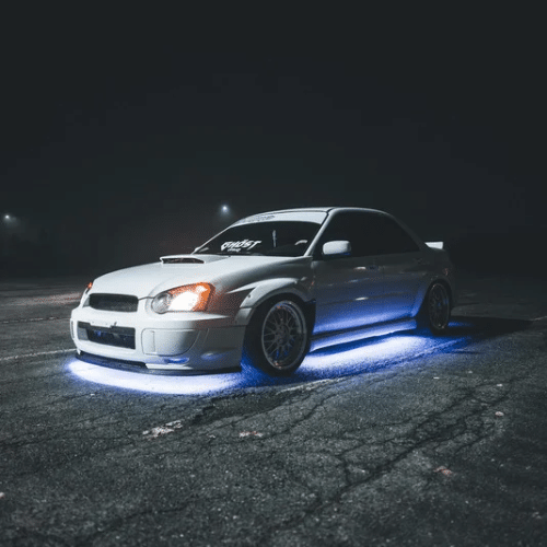 White sports car with blue underglow lights parked on an empty lot at night in South Jordan, with faintly visible fog and distant lights in the background—a striking scene that hints at expert Subaru maintenance and attention to detail.