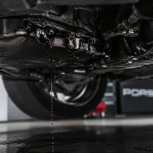 A close-up view of a car's undercarriage with oil visibly leaking and dripping onto the floor below.