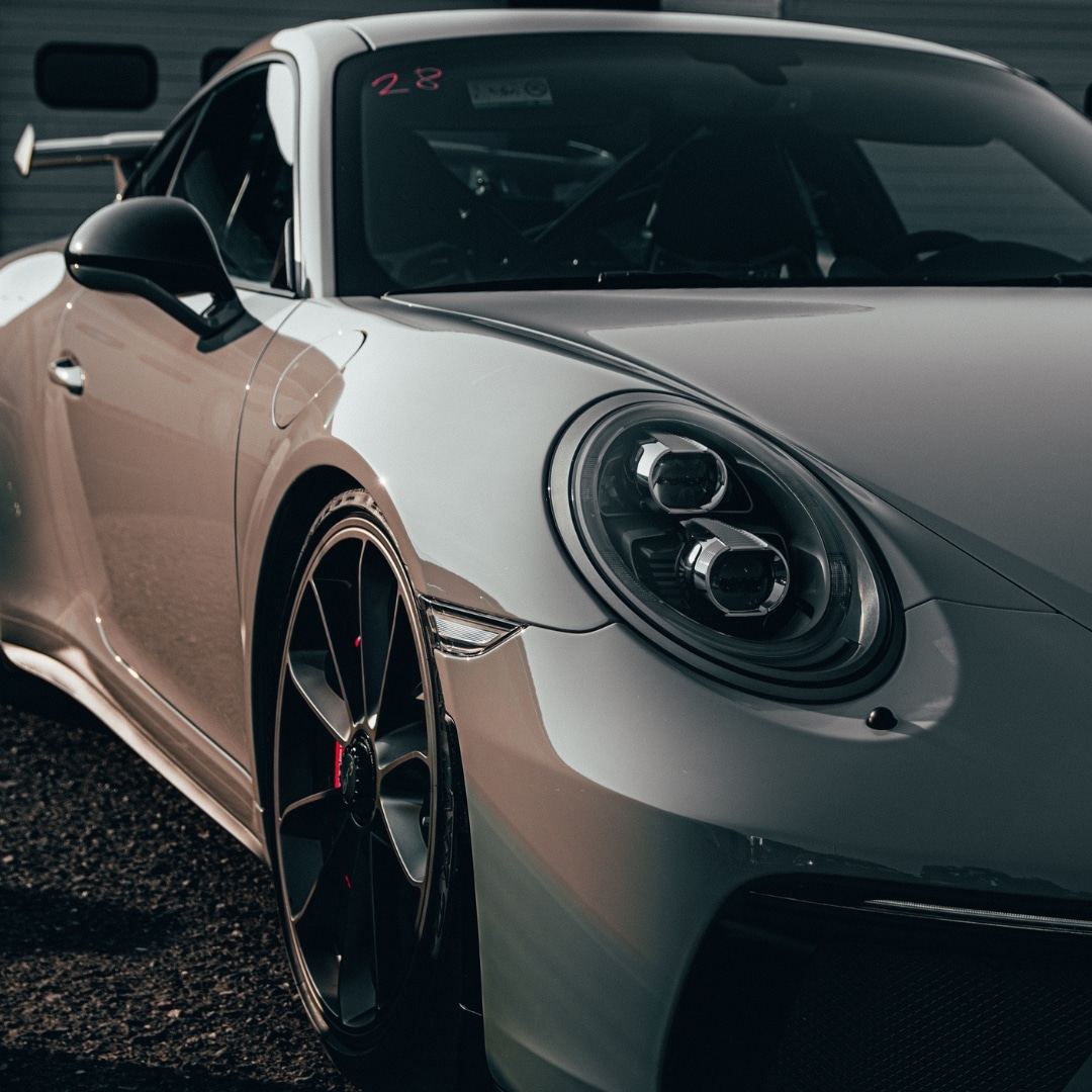 Close-up of the front left side of a silver sports car parked on asphalt, showing the headlight, wheel, and part of the windshield.