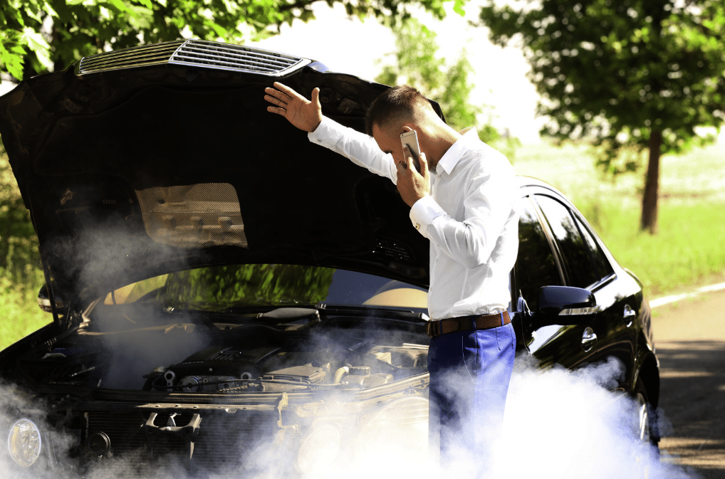 A man stands by his smoking car with the hood open, talking on his phone, appearing to seek help for a car breakdown after his car engine overheats on a sunny day.