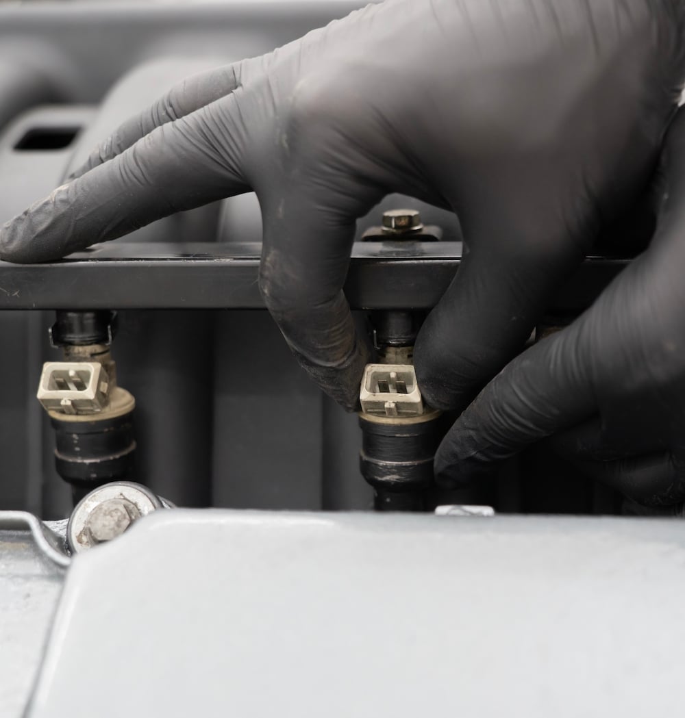 A person wearing black gloves is working on a car engine, adjusting a component connected to fuel injectors as part of a Fuel System Service to enhance vehicle performance.
