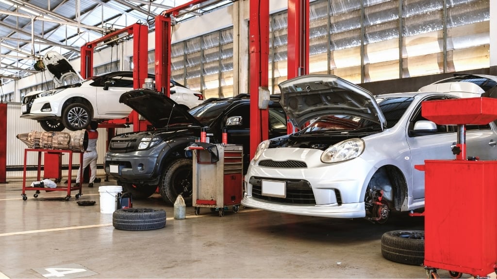 Three cars in an auto repair shop, each on a hydraulic lift with their hoods open; tools and tires are scattered around the work area.
