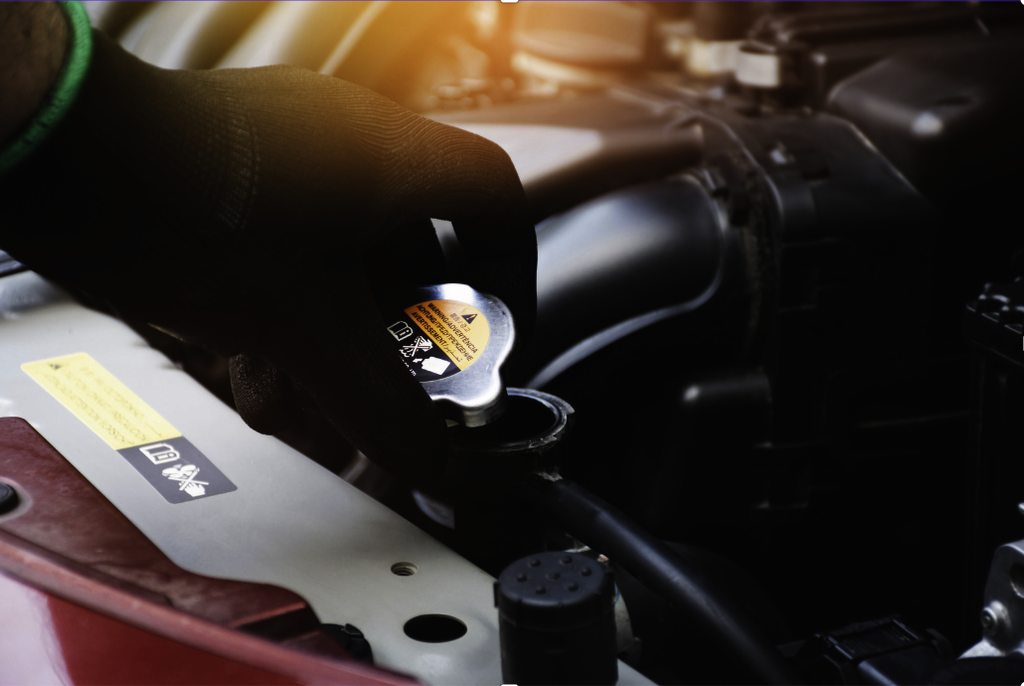 A gloved hand is removing a radiator cap under the hood of a car, with engine components visible in the background—an important step when addressing issues such as car overheating or deciding what to do if your engine overheats.