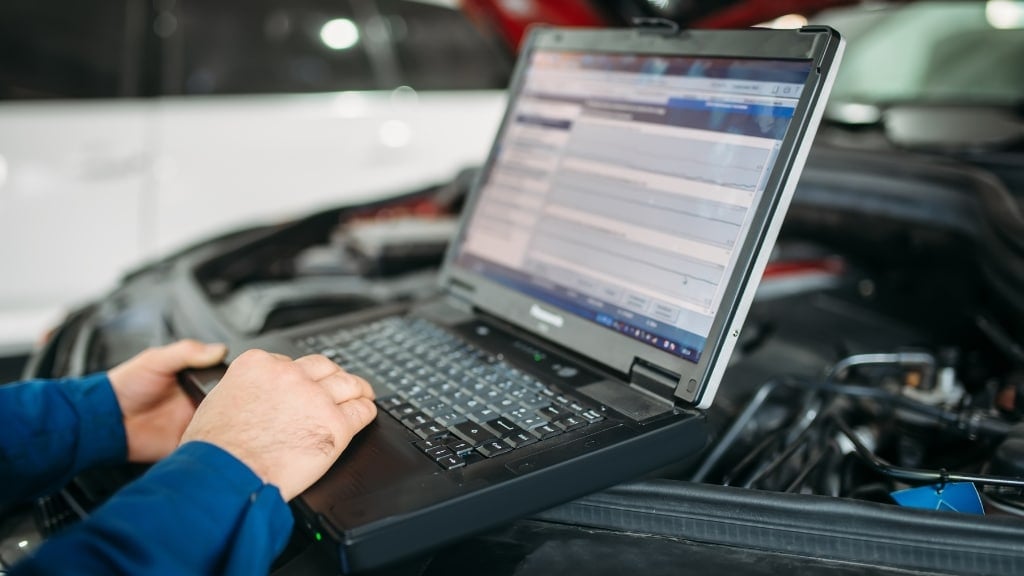 A person uses a laptop computer to run diagnostic software on a vehicle with its hood open.